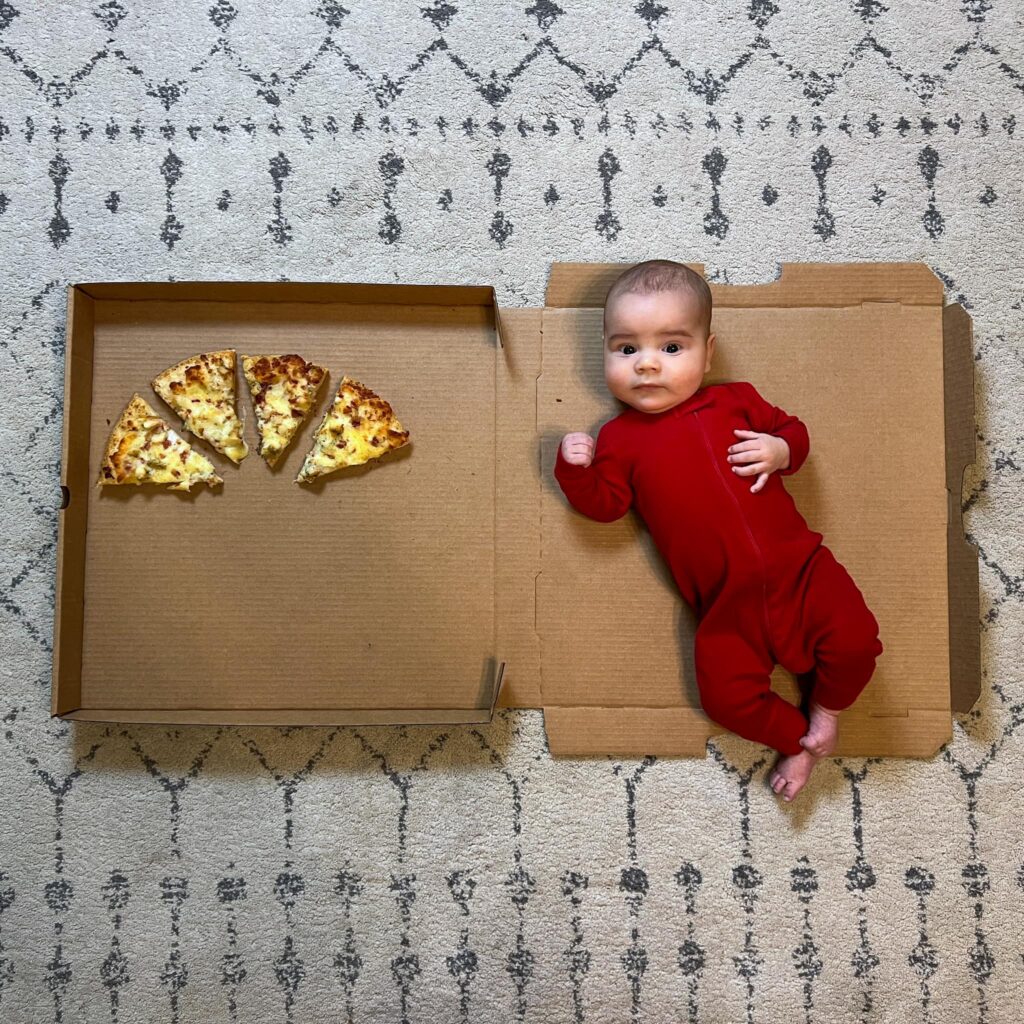 photo shows a baby in a red onesie in a pizza box