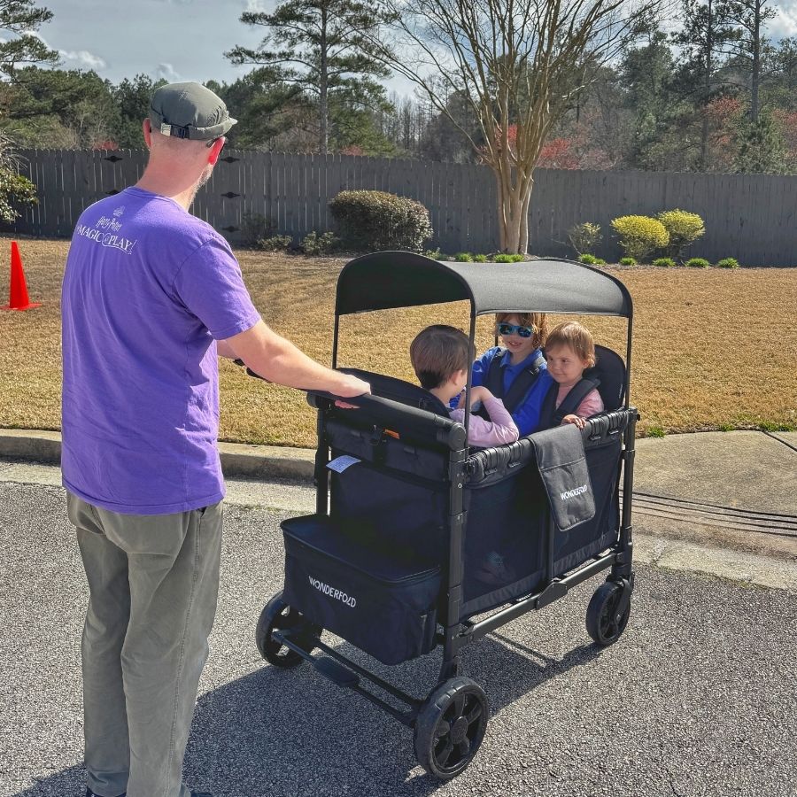 image shows a man pushing three children in a wonderfold wagon